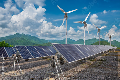 Renewable energy concept. Solar panels with wind turbines against mountanis landscape against blue sky with clouds