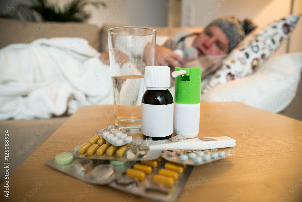 Sick man wearing scarf lying on couch at home under a blanket checking temperature. Living room with table full of medicine and pills. Man with running nose and cough. 