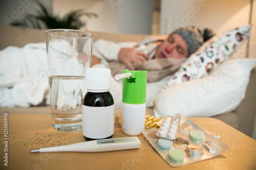 Sick man wearing scarf lying on couch at home under a blanket checking temperature. Living room with table full of medicine and pills. Man with running nose and cough. 