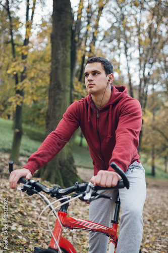 Autumn outdoors. Young serious handsome guy with earphones standing with bicycle in park, holding a cell phone and picking music tracks.