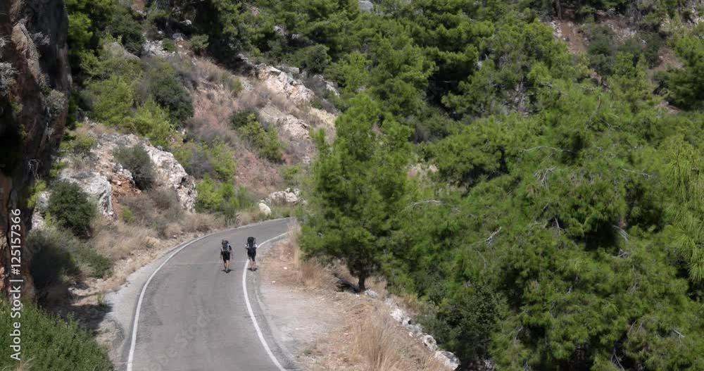 Two Young Tourist Hikes With Backpacks Going by the Road in the Mountains Two Young Tourist Hikes With Backpacks Going by the Road in the Mountains
