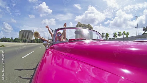 Happy people driving through the Havana city in the pink cabriolet car.Camera fixed at the car, moving camera.Many holes on the roads cause shaking of footage.