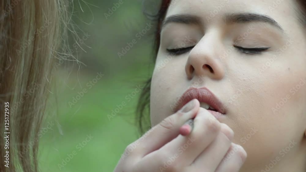 Makeup artist applies lipstick perfectly on a young woman, outdoor scene Makeup artist applies lipstick perfectly on a young woman, outdoor scene