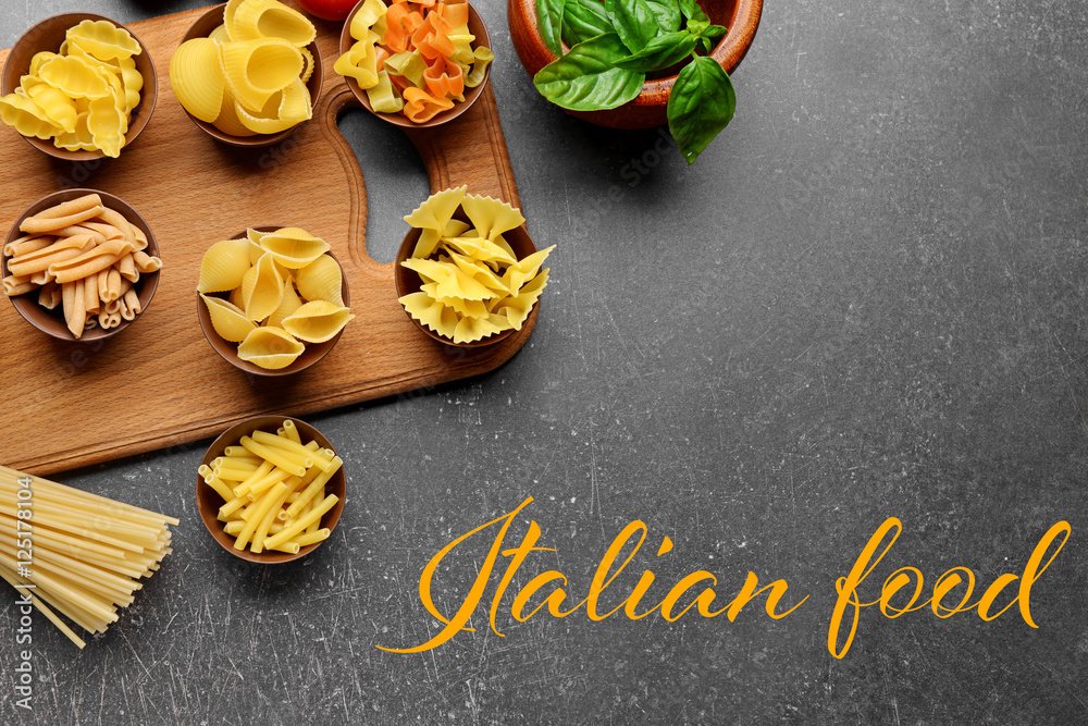 Bowls with different pasta on wooden cutting board. Text ITALIAN FOOD on gray background. Traditional meal concept.