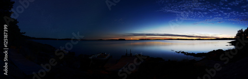 Panorama of a lake at dusk and after nightfall. On the right side sky and lake are illuminated by the sunset. On the left side stars are visible. The foreground is the dark silhouette of the coastline