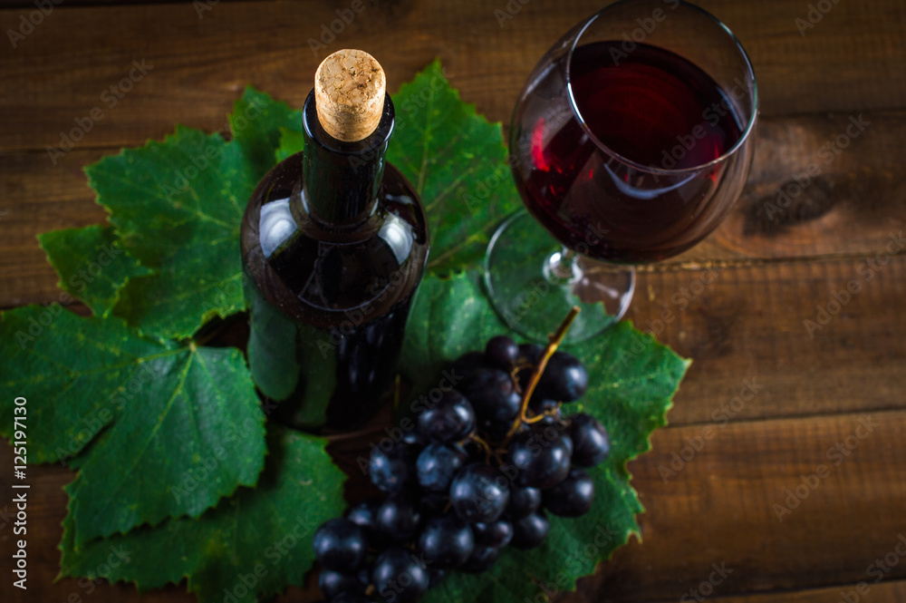 Bottle of red wine, glass. Bunch of grapes on leaves from the grape. On a wooden background.