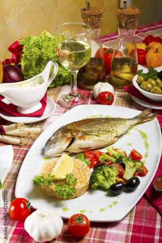 Fried sea bream on a plate with a side dish of broccoli, cherry tomatoes, rice, olives and lemon served beside fresh vegetables, sauce, olive oil and white wine.