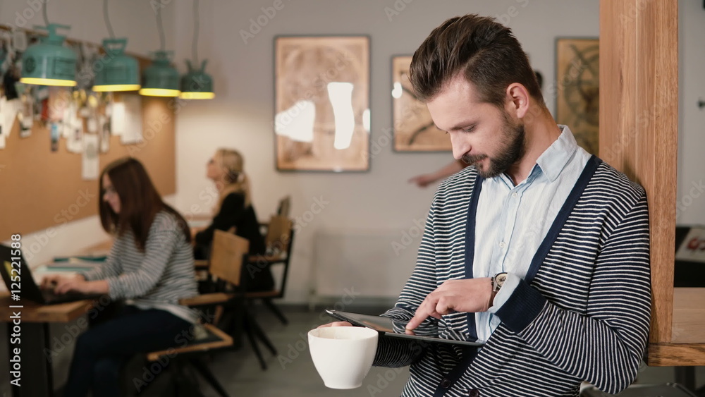 young handsome bearded man uses touchscreen tablet and drinking coffee in the modern startup office. young handsome bearded man uses touchscreen tablet and drinking coffee in the modern startup office.
