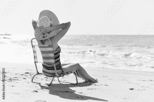 Black and white back side view of happy woman on a tropical beach sitting in chair, sunny background outdoors