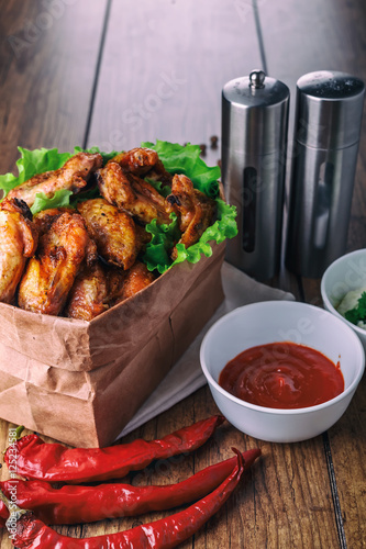 delicious grilled chicken wings with garlic and tomato sauce with lettuce in food paper bag on wooden rustic background top view, witi copyspace photo