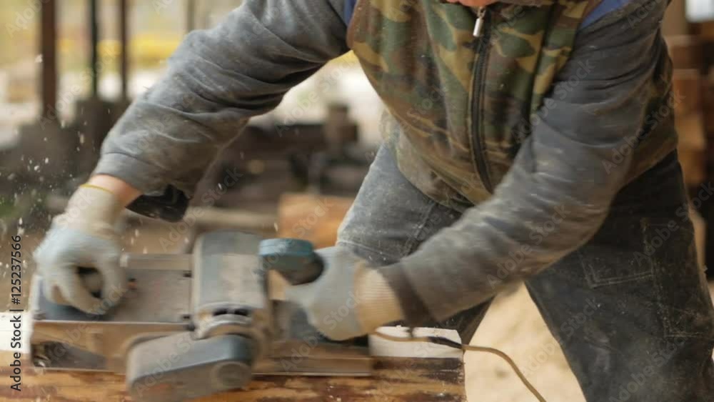 Construction worker planing a piece of wood for a building project. Construction grinds electric hand plane. Against the background of the factory premises, slow motion Construction worker planing a piece of wood for a building project. Construction grinds electric hand plane. Against the background of the factory premises, slow motion