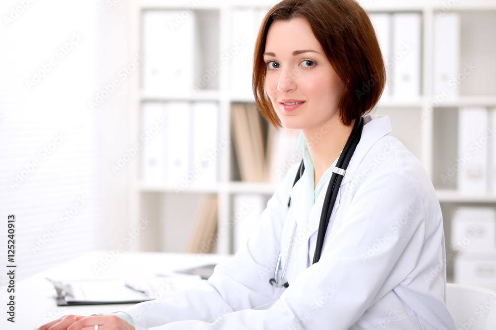 Young brunette female doctor sitting at the table and working at hospital office. Health care, insurance and help concept. Physician ready to examine patient Young brunette female doctor sitting at the table and working at hospital office. Health care, insurance and help concept. Physician ready to examine patient