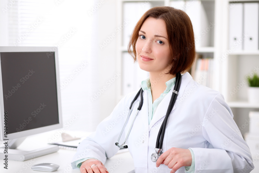 Young brunette female doctor sitting at a desk and working on the computer at the hospital office.  Health care, insurance and help concept. Physician ready to examine patient