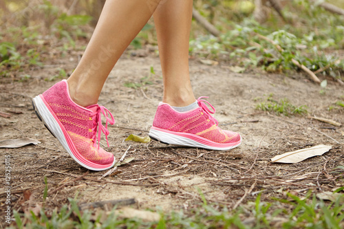 Sports and adventure concept. Close up shot of female legs wearing pink running shoes in forest while exercising in summer nature. Young Caucasian woman athlete walking or hiking on trail in wood