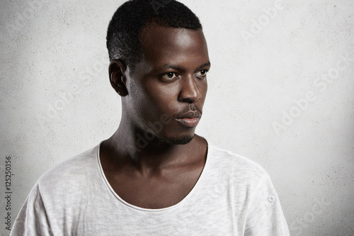 Side view portrait of pensive African man wearing white T-shirt with low neckline looking ahead of him with serious and confident expression, posing isolated against white concrete wall background