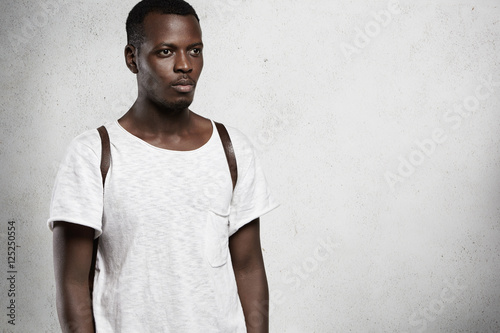 Style design and fashion. Studio shot of serious good-looking African student wearing leather backpack and white stylish T-shirt standing against blank wall background with copy space for your content