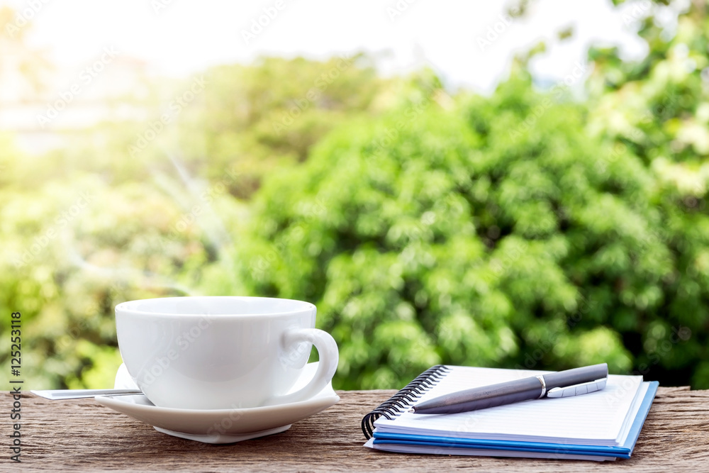 Hot coffee cup with a notebook and a pen on the old wooden floor with colorful green nature background. Hot coffee cup with a notebook and a pen on the old wooden floor with colorful green nature background.