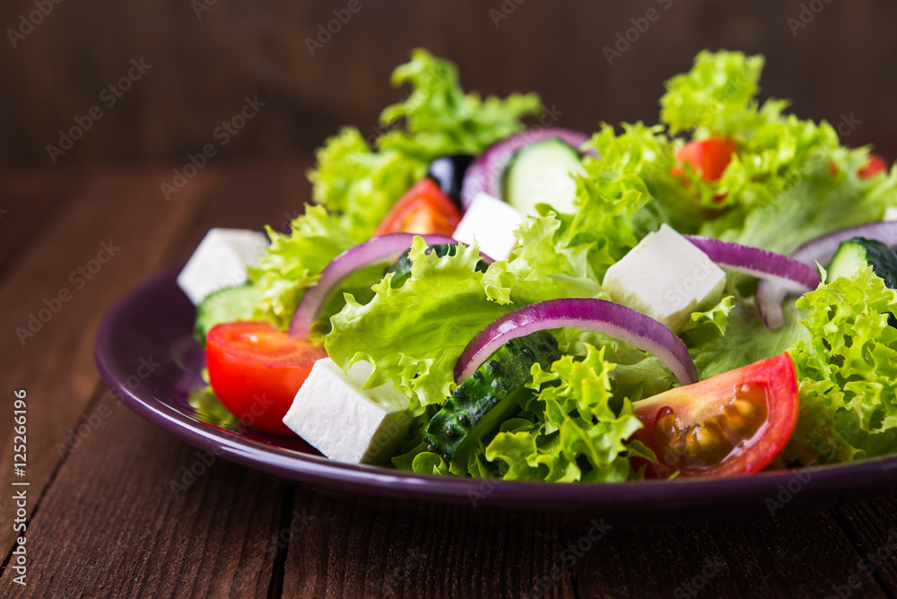 Greek salad (lettuce, tomatoes, feta cheese, cucumbers, black olives, purple onion) on dark wooden background close up. Healthy food. Greek salad (lettuce, tomatoes, feta cheese, cucumbers, black olives, purple onion) on dark wooden background close up. Healthy food.