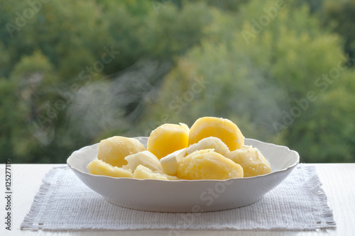 A white plate is a boiled potato. The potatoes are crisp. On potatoes the steam. The plate is on a white napkin on the windowsill.