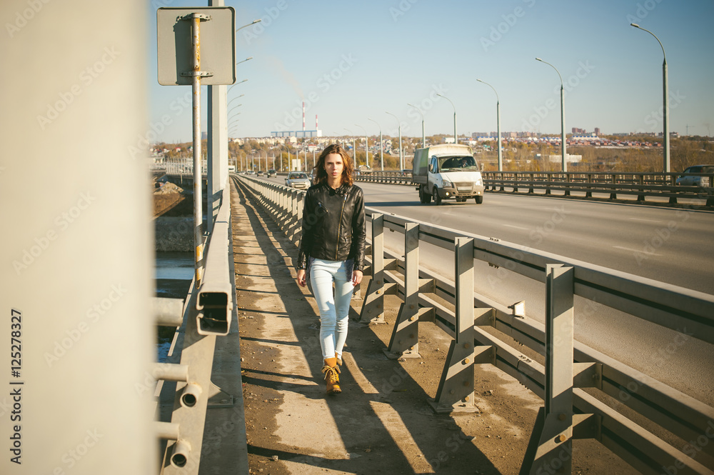 Naklejka premium girl goes on road bridge. lonely young woman in black Leather jacket autumn, walking over the bridge which goes transport.