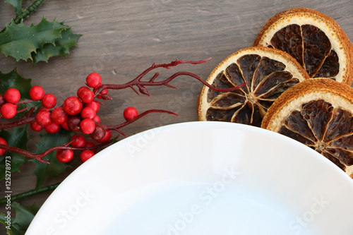Traditional christmas (Xmas) festive decoration background with white plate on wooden background with dried orange and holly branch