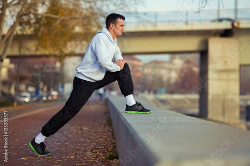 Side view of an attractive young man exercising outdoors. He is doing stretching exercise for his legs and looking away.