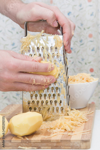 Obraz na płótnie Man's hands with a grater grate the potatoes on the wooden board for potato pancakes in the kitchen