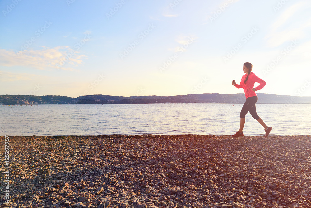 Young fitness woman runner wearing orange jacket and black pants running on stony beach near sea, working out outdoors, listening to music.
