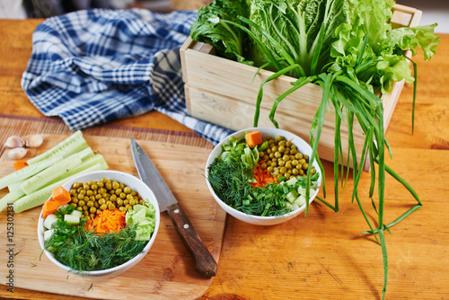 Vegetable mix in bowls with green peas, cucumbers, carrots, lettuce and dill, standing on a wooden table