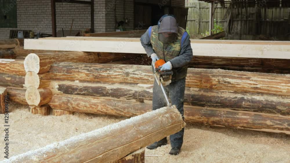 Man makes curly cutting wood Chainsaw. The log will be part of the future of the wooden house. Protective face mask on the face of the builder and a lot of sawdust. Man makes curly cutting wood Chainsaw. The log will be part of the future of the wooden house. Protective face mask on the face of the builder and a lot of sawdust.