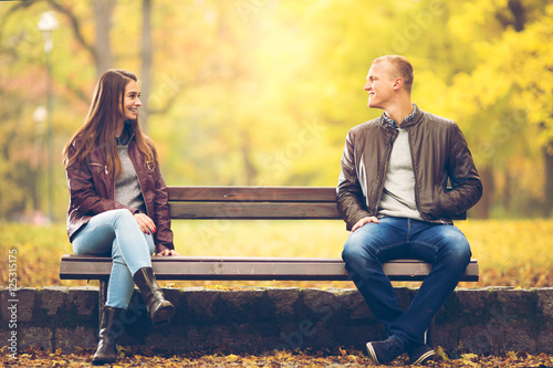 Young couple is sitting on a bench in a park on a beautiful Autumn day. They are talking to each other