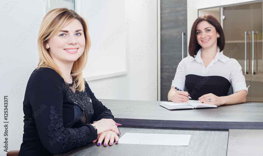 Two women sitting at a table in the office. Blonde and brunette on job interview, or meeting. Two women sitting at a table in the office. Blonde and brunette on job interview, or meeting.
