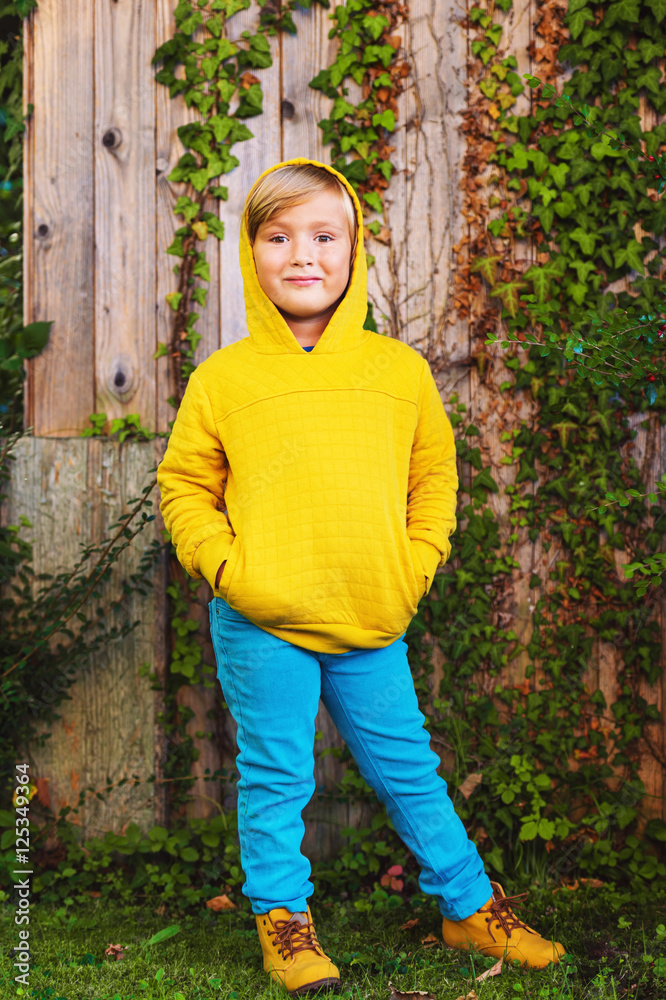 Fototapeta premium Outdoor portrait of adorable little 5 year old blond boy playing in the park, wearing yellow sweatshirt with hood, shoes and blue trousers