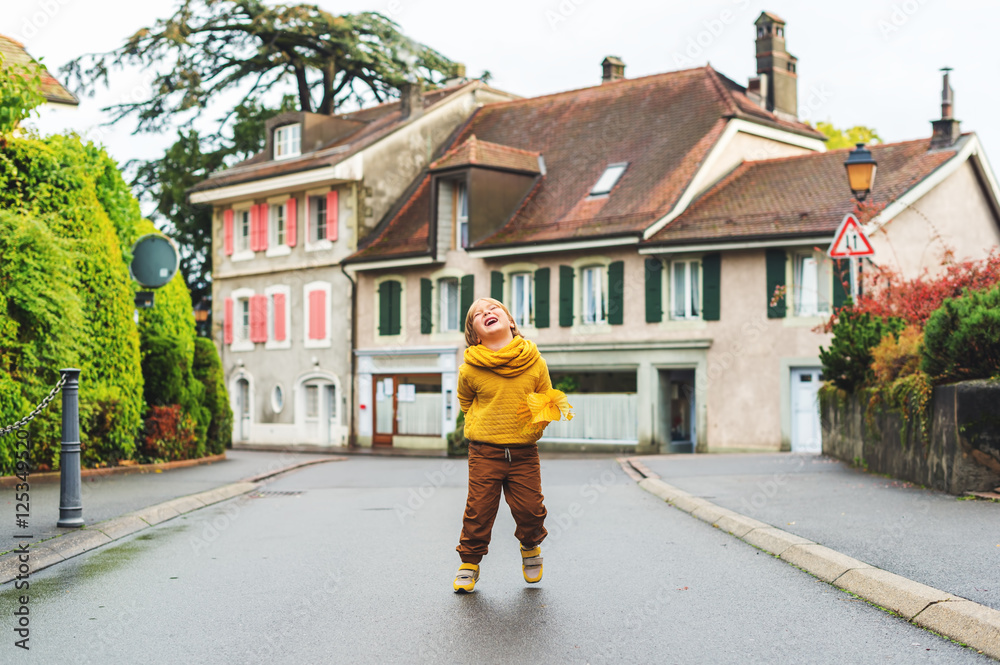 Outdoor fashion portrait of adorable 5-6 year old little blond boy in the street, wearing brown trousers, yellow sweatshirt, scarf and trendy sneakers