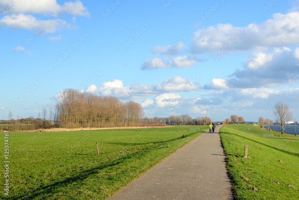 Bike and walking path over an embankment along a wide river in the Netherlands. It's a sunny day in the winter season and in the background an unknown elderly couple is strolling together.