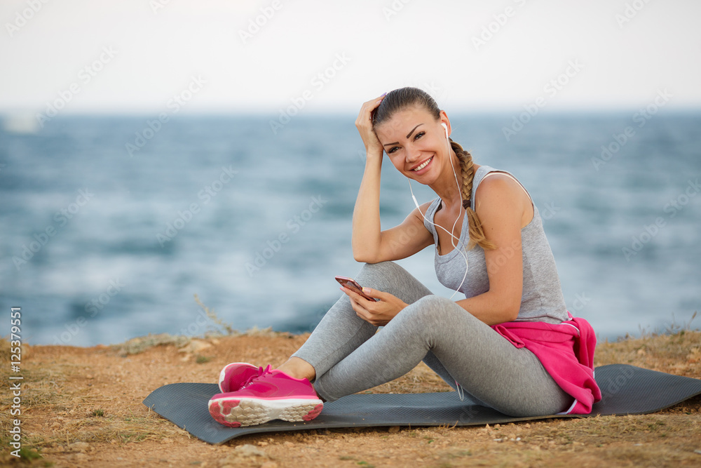 Athletic woman,brunette,hair in a braid,dressed in a grey shirt and grey sweat pants,pink sneakers,sitting on fitness Mat in the background of the sea,listening to music through white headphones