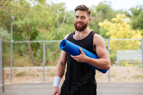 Smiling cheerful fitness man with yoga mat standing outdoors