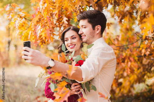 Stylish and romantic caucasian couple smiling and making selfie in beautiful autumn park. Love, relationships, romance, happiness concept. Bouquet in girl's hands.