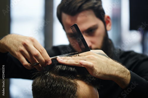 Slika na platnu The Barber a man with a beard in the process of cutting the client a pair of sci