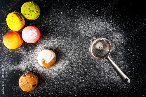 Selection of colorful cakes macarons on a black background, sprinkled with powdered sugar. With a strainer to sift powder and flour. Copy space