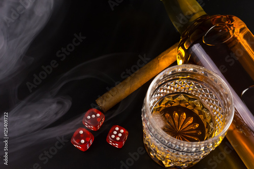A glass of whiskey, a bottle, cigar, smoke and dice on black.