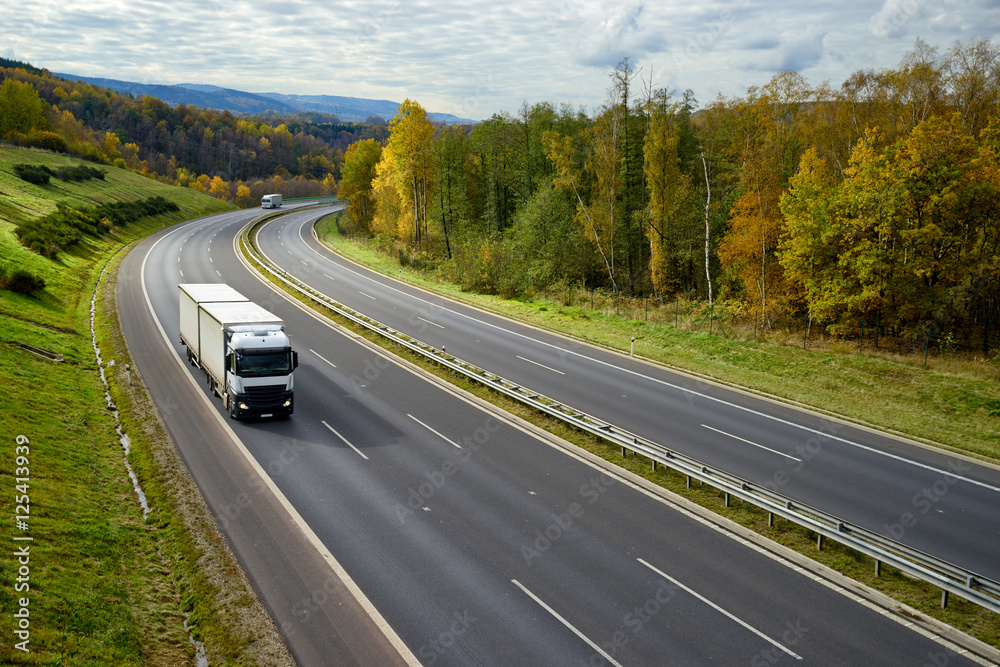 Asphalt highway with oncoming white trucks in the autumn landscape.