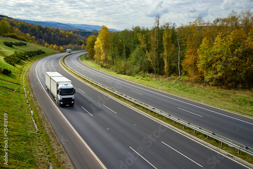 Asphalt highway with oncoming white trucks in the autumn landscape.