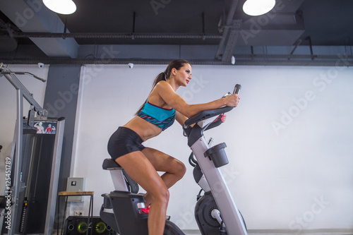 Cute beautiful girl pedaling on a stationary bike at the gym. In the background a gym. Girl dressed in a blue top and black shorts. Training on the simulator