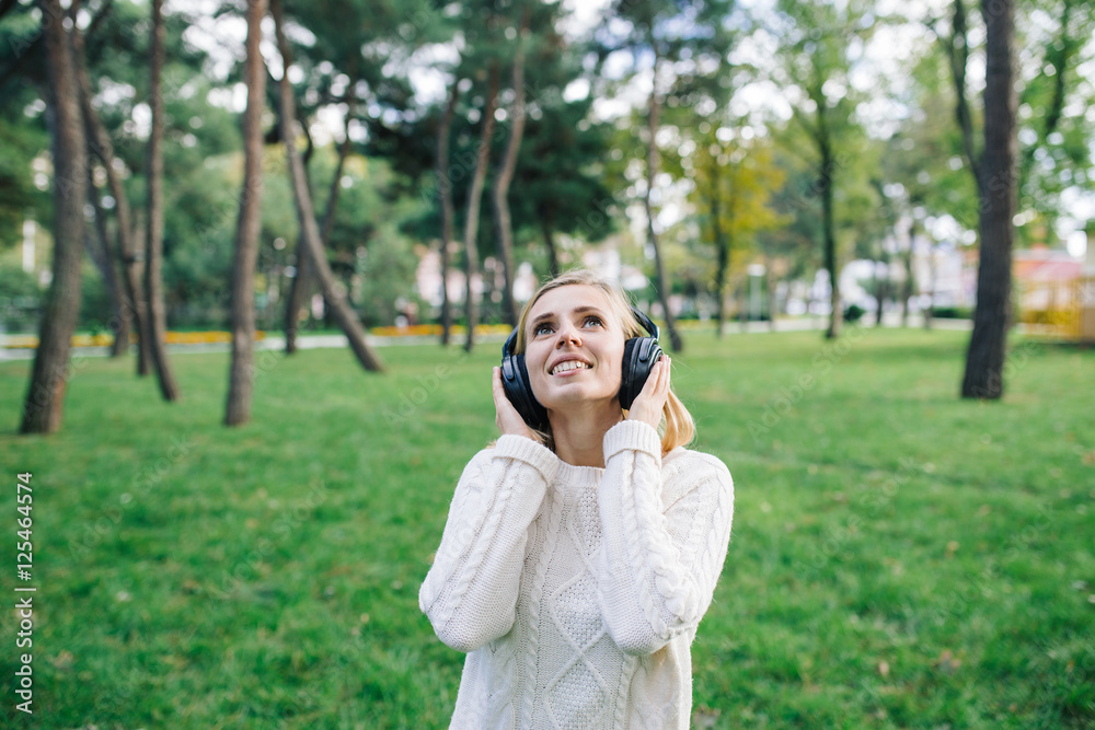 Young beautiful woman enjoying the music outdoor. Happy woman in white sweater with headphones listening to music in the park Young beautiful woman enjoying the music outdoor. Happy woman in white sweater with headphones listening to music in the park