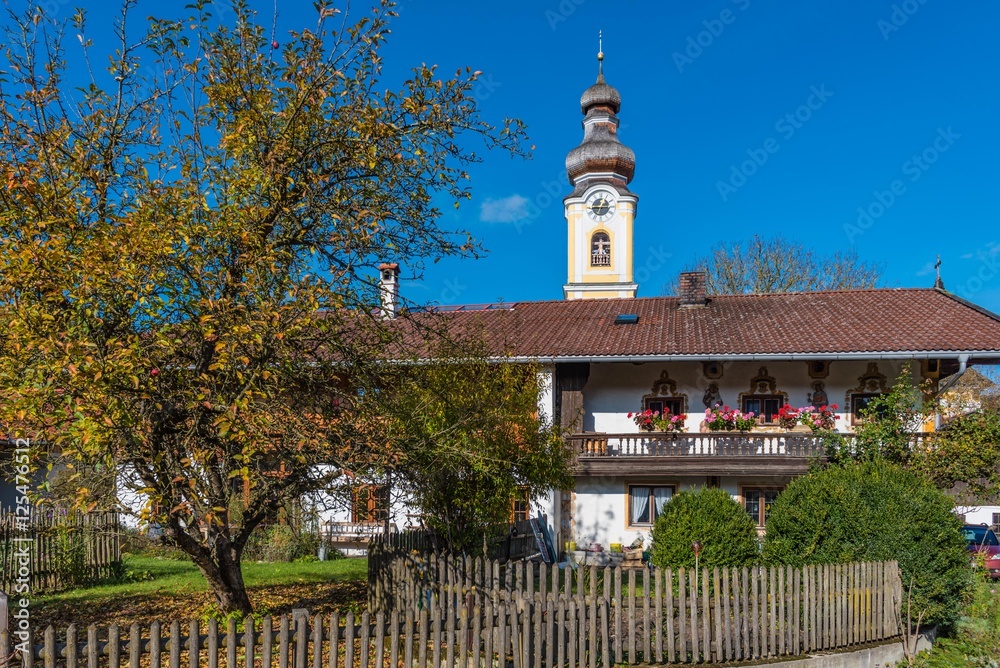Fototapeta premium Bauernhaus mit Holzgartenzaun, Baum und Kirchturm in Bayern