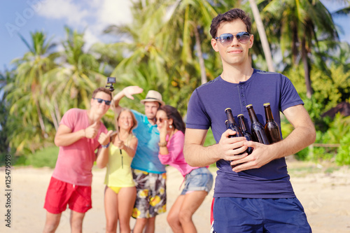 Cheers! Welcome to the beach party. Group of young people having fun together on the sea shore. Young handsome man holding bottles of beer.