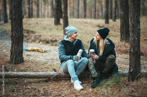 Romantic couple sitting near bonfire, autumn forest background. Young blonde woman and handsome man. Concept - family, togetherness, love, friendship.