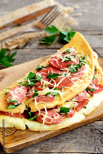 Egg omelette with grated cheese and fried sausages on a board, fork, knife, burlap, fresh parsley leaves on old wooden table. Delicious omelette recipe. Closeup