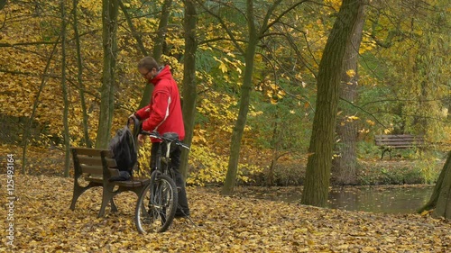 Cyclist Comes Left His Bike Sits Down to Bench Autumn Day Man is Resting Among Golden Trees in Park Ground is Covering With Yellow Leaves Footpath Alley photo